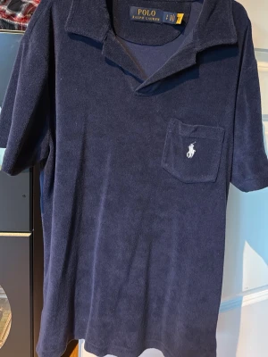 Marinblå frottétröja Polo Ralph Lauren - Marinblå skjorta i mjuk frotté från Polo Ralph Lauren. Skjortan har korta ärmar, klassisk krage och en bröstficka med broderad logga. Perfekt för en avslappnad och stilren look.