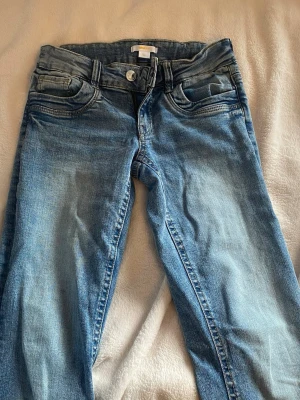 Blå bootcut jeans med slitningar - Säljer ett par ljusblå bootcut jeans med snygga slitningar och klassiska fem fickor. Jeansen har normal midja och dragkedja med knapp. Tillverkade i denim med lätt stretch för skön passform.