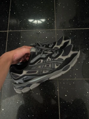 Svarta och grå Asics sneakers - Snygga sneakers från Asics i svart och grått med coola detaljer och meshpaneler för extra andningsförmåga. Skorna har en chunky sula och klassisk snörning. Perfekta för dig som gillar sportig stil och vill ha något bekvämt till vardags.
