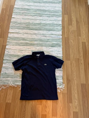 Marinblå pikétröja från Lacoste - Snygg marinblå pikétröja från Lacoste i klassisk passform. Tröjan har korta ärmar, krage med knappar och den ikoniska krokodilloggan på bröstet. Tillverkad i mjuk bomull som känns skön mot huden.