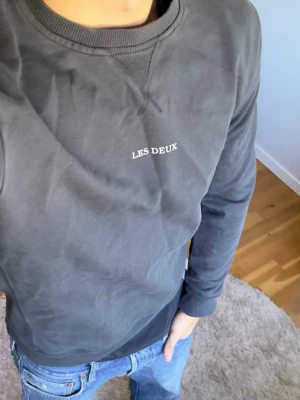 Mörkgrå sweatshirt från Les Deux - Snygg mörkgrå sweatshirt från Les Deux med diskret logga broderad på bröstet. Tröjan har rund halsringning, ribbade muddar vid ärmslut och nederkant samt en relaxed passform. Perfekt till jeans för en clean och enkel look.