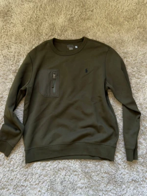 Olivgrön sweatshirt från Polo Ralph Lauren - Snygg olivgrön sweatshirt från Polo Ralph Lauren med rund halsringning och en praktisk bröstficka med dragkedja. Tröjan har ribbade muddar vid ärmslut och nederkant samt den klassiska broderade loggan på bröstet. Perfekt för en avslappnad stil.