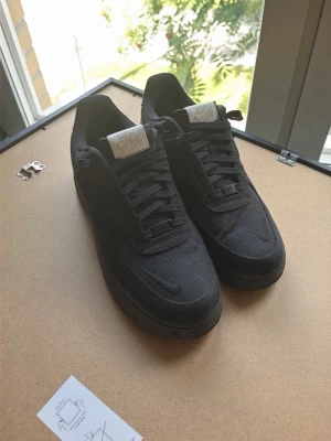 Svarta Nike Air Force 1 sneakers - Säljer ett par klassiska svarta Nike Air Force 1 sneakers i tygmaterial med svart sula och svarta skosnören. Ikonisk Nike Swoosh på sidan och vit Nike Air-logga på plösen. Perfekta för dig som gillar stilrena och tidlösa sneakers.