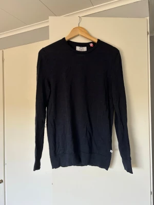 Mörkblå tunn stickad tröja - En stilren svart långärmad tröja i tunn stickad kvalitet ifrån Kronstadt Casual. Tröjan har rund halsringning och ribbade muddar vid ärmslut och nederkant. Perfekt att bära som lager eller för en clean look.