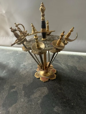 Dekorativt set med miniatyrsvärd i ställ - Säljer ett unikt dekorationsset med flera små svärd i metall, placerade i ett detaljerat ställ av mässing. Svärden har olika former på handtagen och stället har orientaliska mönster i guld och rött. Perfekt som prydnad eller samlarobjekt för dig som gillar annorlunda inredning.