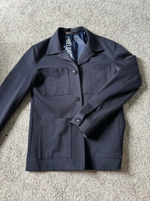 Mörkblå overshirt från Selected homme  - Stilren mörkblå overshirt från Selected med fyra stora fickor framtill och klassisk krage. Jackan har raka ärmar, knäppning med tre knappar och är tillverkad i ett slätt tyg som ger en clean look. Perfekt lager på lager-plagg för en modern stil.