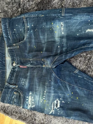 Mörkblå distressed jeans från Dsquared2 - Säljer ett par mörkblå jeans från Dsquared2 med slitningar, färgstänk och en cool, tvättad look. Jeansen har klassisk femficksmodell, låg midja och detaljer som ger en unik streetwear-vibe. Perfekta för dig som gillar en trendig och avslappnad stil.