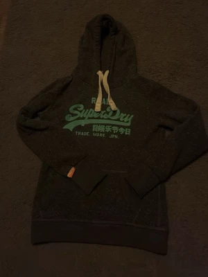 Mörkgrå Superdry hoodie med tryck - Säljer en mörkgrå hoodie från Superdry med stort turkost tryck på bröstet och japanska tecken. Hoodien har vita dragsnören, känguruficka och ribbade muddar. Pris kan såklart diskuteras vid snabb affär hör av er vid minsta fundering.