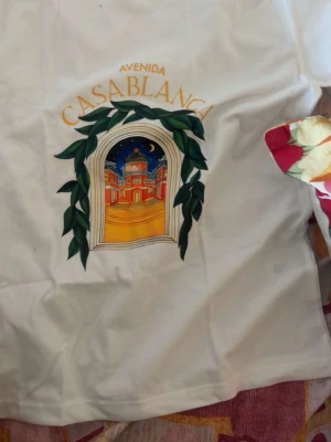 Vit t-shirt med Casablanca-tryck - Vit t-shirt med färgglatt tryck av en nattlig stadsscen och texten 'Avenida Casablanca' på bröstet. Trycket ramas in av gröna blad och en båge, vilket ger en unik och konstnärlig vibe. Perfekt för dig som gillar grafiska motiv och vill sticka ut.