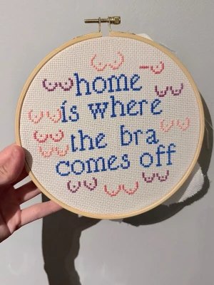 Broderad tavla  - Handgjord broderad tavla med texten 'home is where the bra comes off' i blått garn. Tavlan är dekorerad med små broderade bröst i rosa, lila och rött. Pris är för ram+material+arbetstid. 
