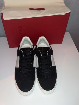 Svarta Valentino Flycrews  - Snygga sneakers från Valentino i svart mocka med vita detaljer och vit sula. Klassisk låg modell med snörning och coola nitar på sidan av sulan. Perfekta för dig som vill ha en stilren men ändå edgy look. Allt og medföljer, nyskick