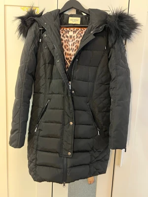Svart lång dunjacka med fuskpäls - Snygg svart lång dunjacka med avtagbar huva och fuskpälsdetalj. Jackan har dragkedjor på fickorna och leopardmönstrat innerfoder. Perfekt för kalla vinterdagar och har en modern, quiltad design.