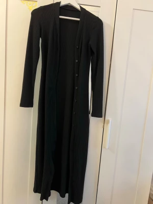 Lång svart ribbad kofta med knappar - Snygg lång svart kofta med ribbad struktur och knappar hela vägen framtill. Koftan har lång ärm och en smal, figurnära passform som ger en stilren look. Perfekt att slänga över en outfit för extra lager och stil.