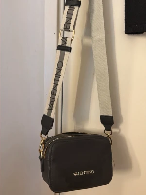 Svart axelväska från Valentino - Snygg svart axelväska från Valentino med guldfärgade detaljer och två olika axelband – ett bredare beige med logga och ett smalare svart. Väskan har en rektangulär form och dragkedja upptill. Perfekt för att bära det viktigaste med stil.        