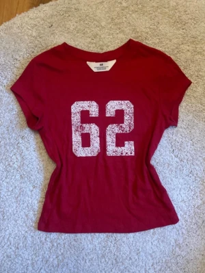 Röd t-shirt - Snygg röd t-shirt från H&M med stort vitt tryck av siffran 62 på framsidan. T-shirten har rund halsringning och korta ärmar. Perfekt för en sportig och avslappnad stil.
