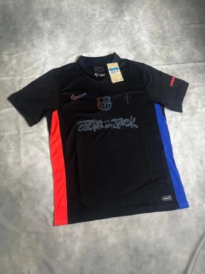 Svart FC Barcelona matchtröja Nike - Snygg svart FC Barcelona matchtröja från Nike med klubbmärke på bröstet, röda och blå sidopaneler samt tryck på bröst och rygg. Kortärmad modell i lätt och ventilerande material, perfekt för fotboll eller träning.