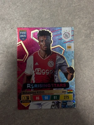 Panini FIFA 365 Mohammed Kudus kort - Samlarbild från Panini FIFA 365-serien med Mohammed Kudus i Ajax matchtröja. Kortet har färgstark design med holografiska detaljer och texten 'Rising Stars'. Siffror och statistik syns på framsidan. Baksidan har Panini-logga och fotbollsplan.
