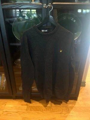 Mörkgrå stickad tröja Lyle & Scott - Mörkgrå stickad tröja från Lyle & Scott med rund halsringning och klassisk gul logga broderad på bröstet. Tröjan har långa ärmar och ribbade muddar vid ärmslut och nederkant. Perfekt för lager-på-lager-stil. Går att Pruta!
