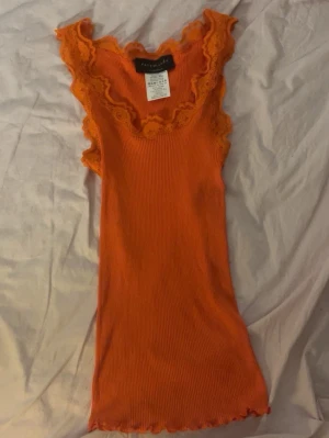 Orange ribbad linnetopp från Rosemunde - Säljer en orange ribbad linnetopp från Rosemunde med spetsdetaljer runt halsringningen och ärmhålen. Toppen är ärmlös och har en figurnära passform. Perfekt för att addera färg till din outfit!