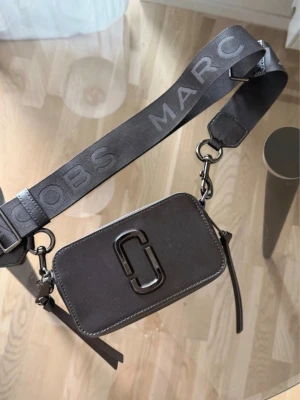 Svart axelväska Marc Jacobs - Snygg svart axelväska från Marc Jacobs med bred justerbar axelrem där märkesnamnet står tryckt. Väskan har en rektangulär form, metallogo framtill och dragkedja.