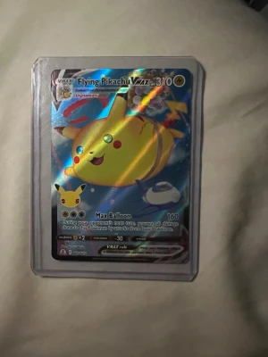 Pokémonkort Flying Pikachu VMAX - Säljer ett Pokémonkort Flying Pikachu VMAX med holografisk yta. Kortet har färgglad design med Pikachu som flyger bland ballonger och är inplastat i ett skyddsfodral. Perfekt för samlare eller Pokémonfans.