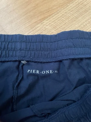 Mörkblå pyjamasbyxor från Pier One, storlek XL - Mörkblå pyjamasbyxor från Pier One i storlek XL. Byxorna har resår i midjan med dragsko och raka ben. Perfekta för att sova i eller chill dagar hemma. Materialet som är bomull känns mjukt och lätt, och de har en enkel, stilren design utan tryck eller mönster.