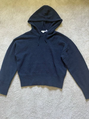 Mörkblå stickad hoodie - Mysig mörkblå hoodie med huva och snören. Tröjan är stickad i ett mjukt material och har långa ärmar samt bred mudd vid midjan. 