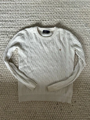Vit kabelstickad tröja Morris Lady - Kabelstickad vit tröja från Morris Lady med rund halsringning och diskret broderad logga på bröstet. Tröjan har långa ärmar och ribbade muddar vid ärmslut och nederkant. Perfekt för en klassisk och stilren look.