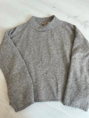 Grå stickad tröja från H&M - Mysig grå stickad tröja från H&M med rund halsringning och ribbade muddar. Tröjan har en enkel och stilren design som passar perfekt till lager på lager och är superbekväm för kalla dagar.