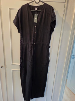 Svart jumpsuit med knappar strl M  - Stilren svart jumpsuit med korta ärmar och knappar framtill. Har en avslappnad passform, markerad midja med knytband i sidan och V-ringning. Tillverkad i ett mjukt, tunt tyg som ger en skön känsla. Perfekt för dig som gillar enkel och trendig stil. Oanvänd. 