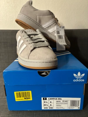 Adidas Campus 00s grå sneakers NYA - Helt nya Adidas Campus 00s sneakers i ljusgrå mocka med klassiska vita ränder på sidorna och en gummisula i beige. Skorna har rund tå, platt sula och breda snören för en retro vibe. Perfekt för dig som gillar stilrena och tidlösa sneakers med skön passform.