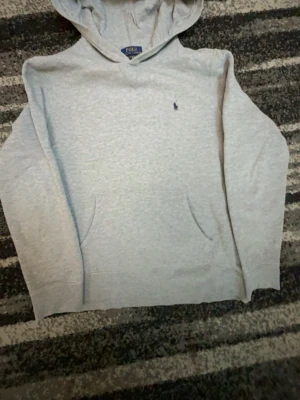 Grå hoodie från Polo Ralph Lauren - Snygg ljusgrå hoodie från Polo Ralph Lauren med klassisk broderad logga på bröstet. Hoodien har huva, känguruficka och ribbade muddar. Perfekt för en chill och stilren look. Materialet känns mjukt och bekvämt, troligen bomullsblandning.