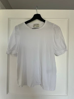 Vit topp med puffärm från AWARE/VERO MODA - Säljer en stilren vit topp från AWARE med puffärmar och rund halsringning. Toppen är gjord i mjuk bomull och har en enkel, clean look som passar till det mesta. Perfekt för dig som gillar minimalistisk stil och vill ha något bekvämt men ändå trendigt.