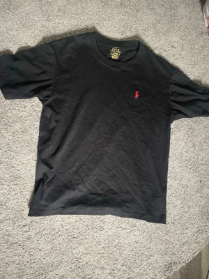 Svart t-shirt från Polo Ralph Lauren - Klassisk svart t-shirt från Polo Ralph Lauren med röd broderad logga på bröstet. Modellen är custom slim fit och har korta ärmar samt rund halsringning. Tillverkad i mjuk bomull som känns skön mot huden. Perfekt till jeans eller shorts.