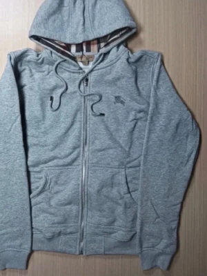 Burburry hoodie - Säljer en stilren grå hoodie med dragkedja och huva. Tröjan har dragsko i huvan, två fickor framtill och ribbade muddar vid ärmar och nederkant. Perfekt för chill dagar eller när du vill ha en enkel och snygg look.