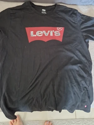 Svart Levi's t-shirt med logga - Klassisk svart t-shirt från Levi's med den ikoniska röda loggan och vit text på bröstet. T-shirten har rund hals och är tillverkad i mjuk bomull. Perfekt för en avslappnad och stilren look.