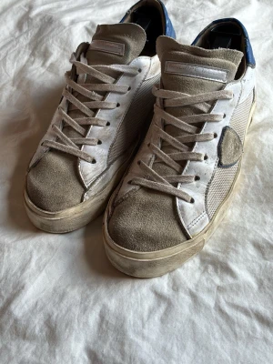 Philippe model skor - Säljer ett par vita och beige sneakers med mockadetaljer och blå häl. Skorna har vita snören, meshpaneler på sidorna och en robust sula. Perfekta för dig som gillar en avslappnad och trendig stil.