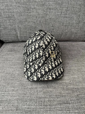 Svartvit Dior keps - Säljer en snygg svartvit keps från Dior med det klassiska Dior-monogrammönstret över hela kepsen. Kepsen har böjd skärm och en guldfärgad Dior-logga på sidan. Materialet ser ut att vara tyg med struktur. Perfekt för dig som gillar exklusiva accessoarer.