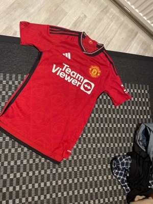 Manchester United matchtröja röd - Säljer en röd Manchester United matchtröja från Adidas med svarta detaljer och klubbmärke på bröstet. Tröjan har TeamViewer-logga framtill, korta ärmar och sportig passform. Perfekt för fotbollsträning eller att bära till vardags.can negotiate for lower price