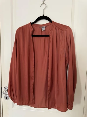 Rostfärgad blus med knappar - Säljer en snygg rostfärgad blus med långa ärmar och knappar framtill. Blusen har en avslappnad passform, rund halsringning utan krage och fina veckade detaljer vid axlar och ärmslut. Materialet är tunt och mjukt, perfekt för lager på lager.
