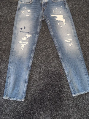 Ljusblå slitna jeans med raka ben - Säljer ett par ljusblå jeans med raka ben och tydliga slitningar och distressed detaljer framtill. Jeansen har klassisk femficksmodell och är tillverkade i denim. Perfekta för en avslappnad och trendig look.
