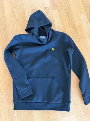 Svart junior hoodie från Lyle & Scott - Svart hoodie från Lyle & Scott med klassisk gul logga på bröstet. Tröjan har huva och en stor magficka framtill. Perfekt för en chill och stilren look. Tillverkad i mjukt material som är skönt att bära hela dagen.
