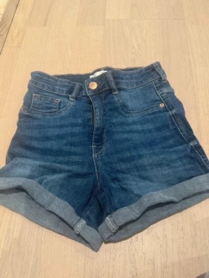Blå denimshorts med upprullad kant - Snygga blå jeansshorts i klassisk modell med upprullad kant. Shortsen har, knapp och dragkedja framtill samt bälteshällor. Perfekta för sommaren och passar till det mesta. Gjorda i mjuk bomullsdenim för skön känsla.