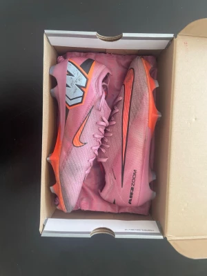 Rosa Nike Air Zoom fotbollsskor - Säljer ett par snygga rosa Nike Air Zoom fotbollsskor med orange och svarta detaljer. Skorna har låg profil, snörning och en lätt ovandel i syntetmaterial för bra bollkontroll. Perfekta för dig som vill sticka ut på planen!
