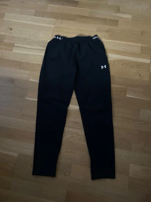 Svarta mjukisbyxor Under Armour - Svarta mjukisbyxor från Under Armour med logga på vänster lår och resår i midjan. Byxorna har smal passform och är perfekta för chill eller träning. Tillverkade i mjukt och stretchigt material för maximal komfort.