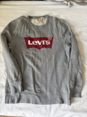 Grå sweatshirt från Levi's - Säljer en grå sweatshirt från Levi's med det klassiska röda logotrycket på bröstet. Tröjan har rund halsringning, långa ärmar och ribbade muddar vid ärmslut och nederkant. Tillverkad i mjukt bomullsmaterial för en skön och avslappnad stil.