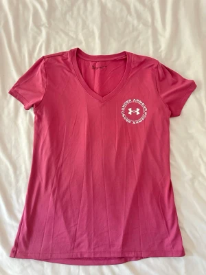 Rosa v-ringad t-shirt från Under Armour - Snygg rosa t-shirt från Under Armour med v-ringning och vit logga på bröstet. Perfekt för träning eller en sportig look!!