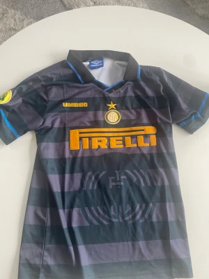 Inter Milan Umbro fotbollströja - Säljer en klassisk Inter Milan fotbollströja från Umbro med svarta och mörkgrå ränder, gula detaljer och Pirelli-logga på bröstet. På ryggen står det 'Ronaldo' och nummer 10 i gult. Tröjan har krage och korta ärmar, perfekt för fotbollsälskare.