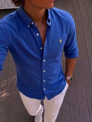 Blå skjorta från Ralph Lauren - Klassisk blå skjorta från Ralph Lauren med button-down krage och vita knappar. Skjortan har lång ärm och den ikoniska gula logotypen broderad på bröstet. Perfekt till jeans eller chinos för en clean och stilren look.