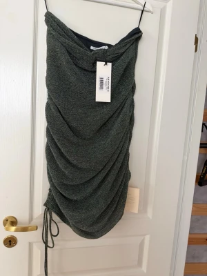 Glittrig grön kortklänning från Adoore - Säljer en glittrig grön kortklänning från Adoore med draperad effekt och snörning i sidan. Klänningen har en asymmetrisk design och är perfekt för dig som vill sticka ut. Materialet är syntet och ger ett snyggt skimmer.